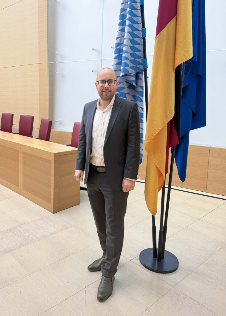 Bild von Felix Locke stehend, vor der bayerischen und deutschen Flagge sowie der Flagge der EU